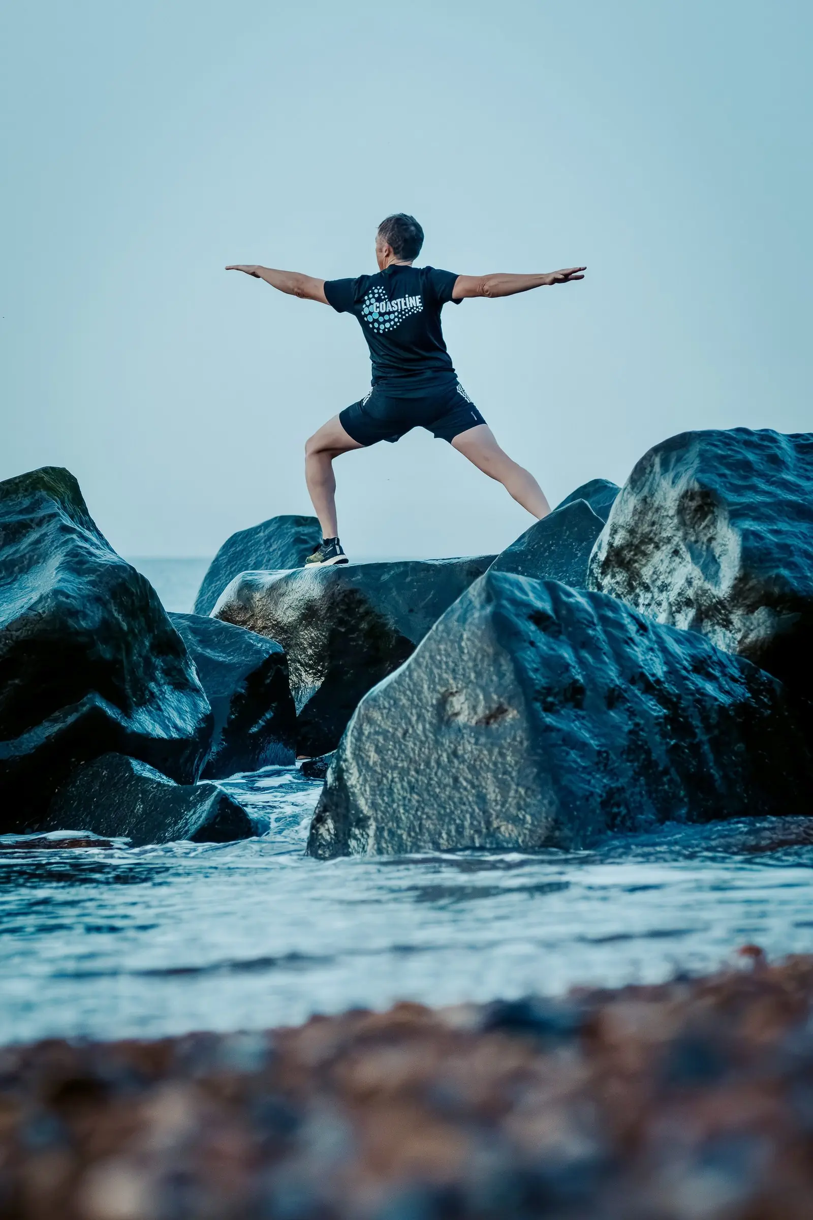 Mark in a wide warrior stance on the rocks, seen from behind, arms out level
