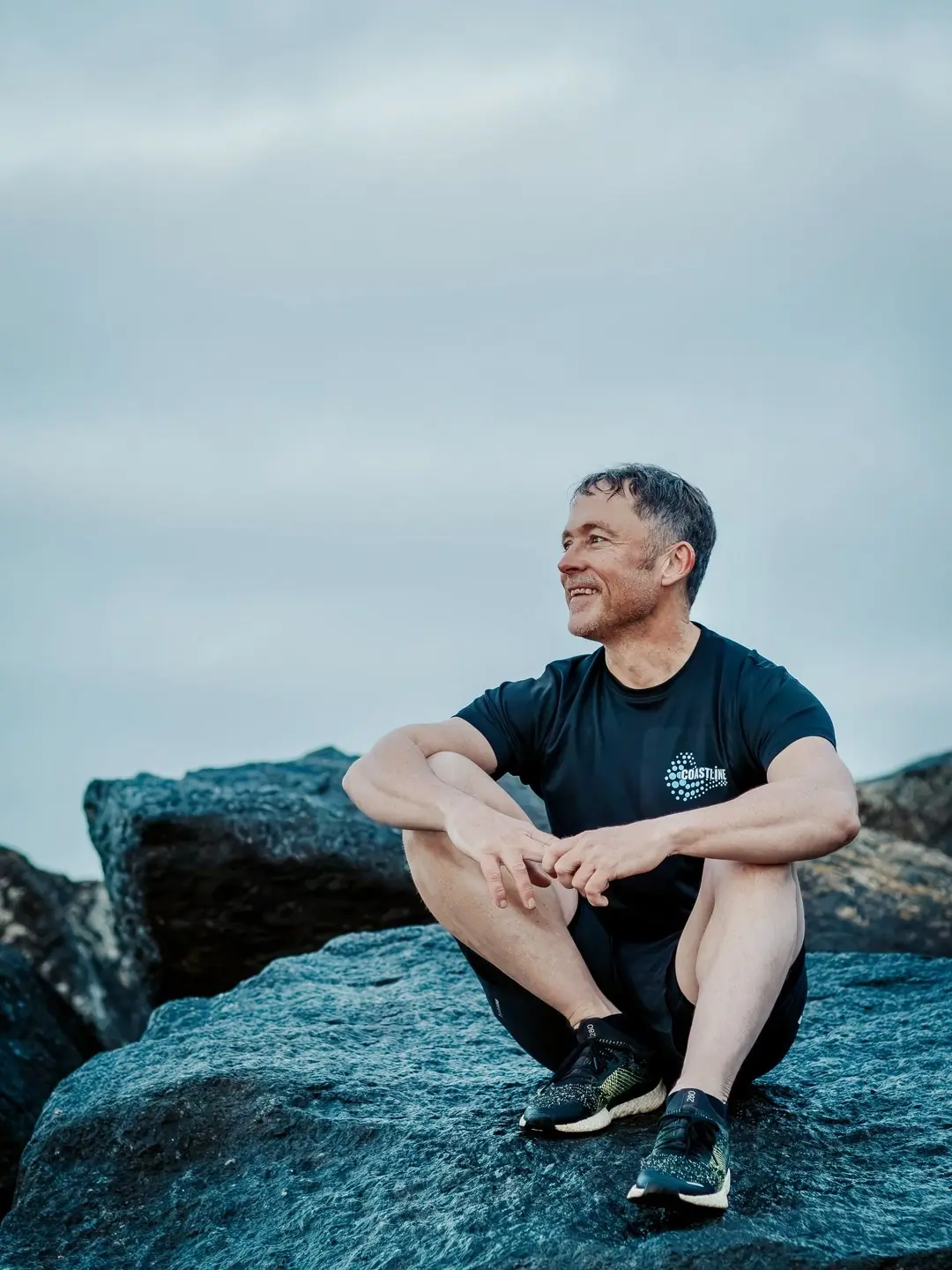 Mark sitting on a rock groyne on Shoreham beach in a Coastline T-shirt, smiling